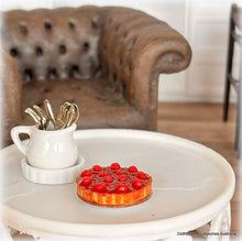 Small round table with a white mug holding gold spoons and a decorative red and orange item on a blurred background of a brown leather sofa.