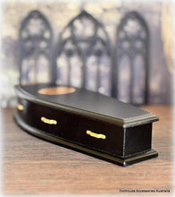 Black miniature coffin with gold handles on a wooden surface, blurred gothic architecture in the background.