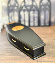 Black coffin with gold accents on a wooden surface, blurred gothic architecture in the background.
