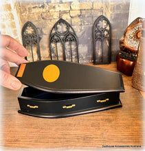 Black coffin-shaped box with a yellow circle on a wooden surface, with decorative elements in the background.