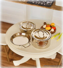 Set of silver pots and pans on a small round table with miniature food items.
