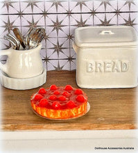 Dollhouse accessories including a pie, bread box, and utensils on a wooden surface with a tiled wall background.