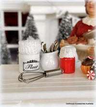 Miniature kitchen scene with flour container, utensils, and a whisk on a white surface.