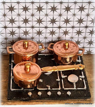Set of miniature copper pots and pans on a stove with a tiled wall background